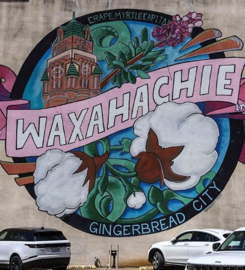 Colorful mural celebrating waxahachie with references to its reputations as "crape myrtle capital" and "gingerbread city," with parked cars in the foreground.