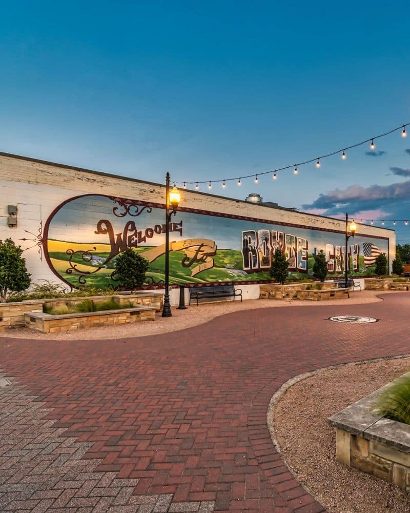 A pedestrian plaza with string lights and benches in front of a building with large mural art during dusk.