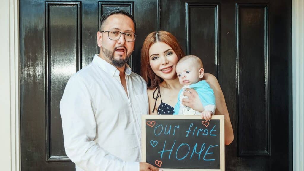 Picture of a young family in front of a door holding a sign that says Our First Home