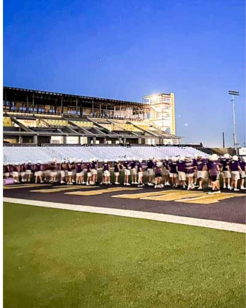 A group of people standing on a sports field with a stadium in the background during dusk.